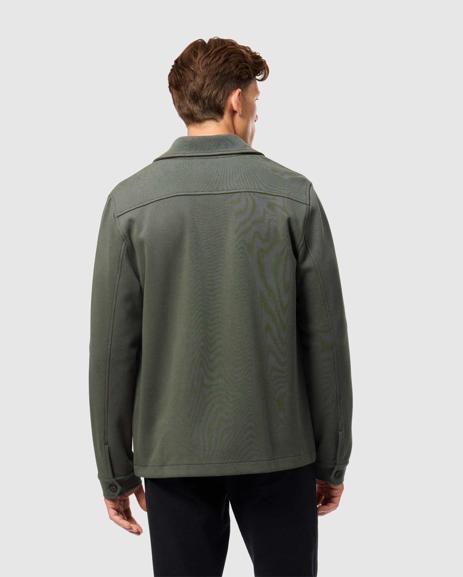 A person with short brown hair faces away from the camera, showcasing an urban vibe in Psycho Bunny's dark green cotton-blend Men's Bedford Shirt Jacket (B6J290B200) paired with black pants against a light gray backdrop.