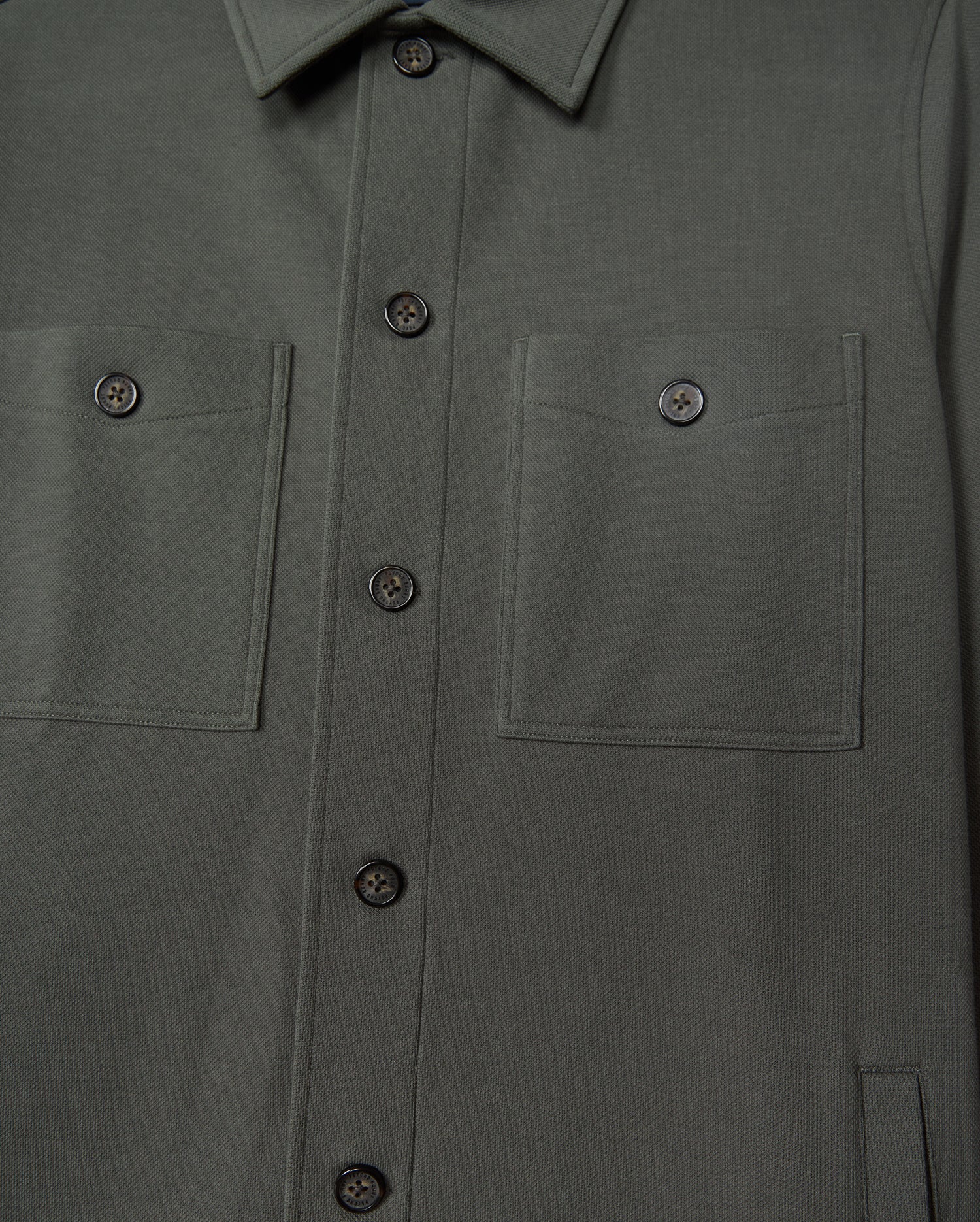 Close-up of the Psycho Bunny Men's Bedford Shirt Jacket (B6J290B200) in dark green cotton-blend. The urban-style jacket highlights two chest pockets, black buttons, neatly stitched seams, and a fold-over collar.