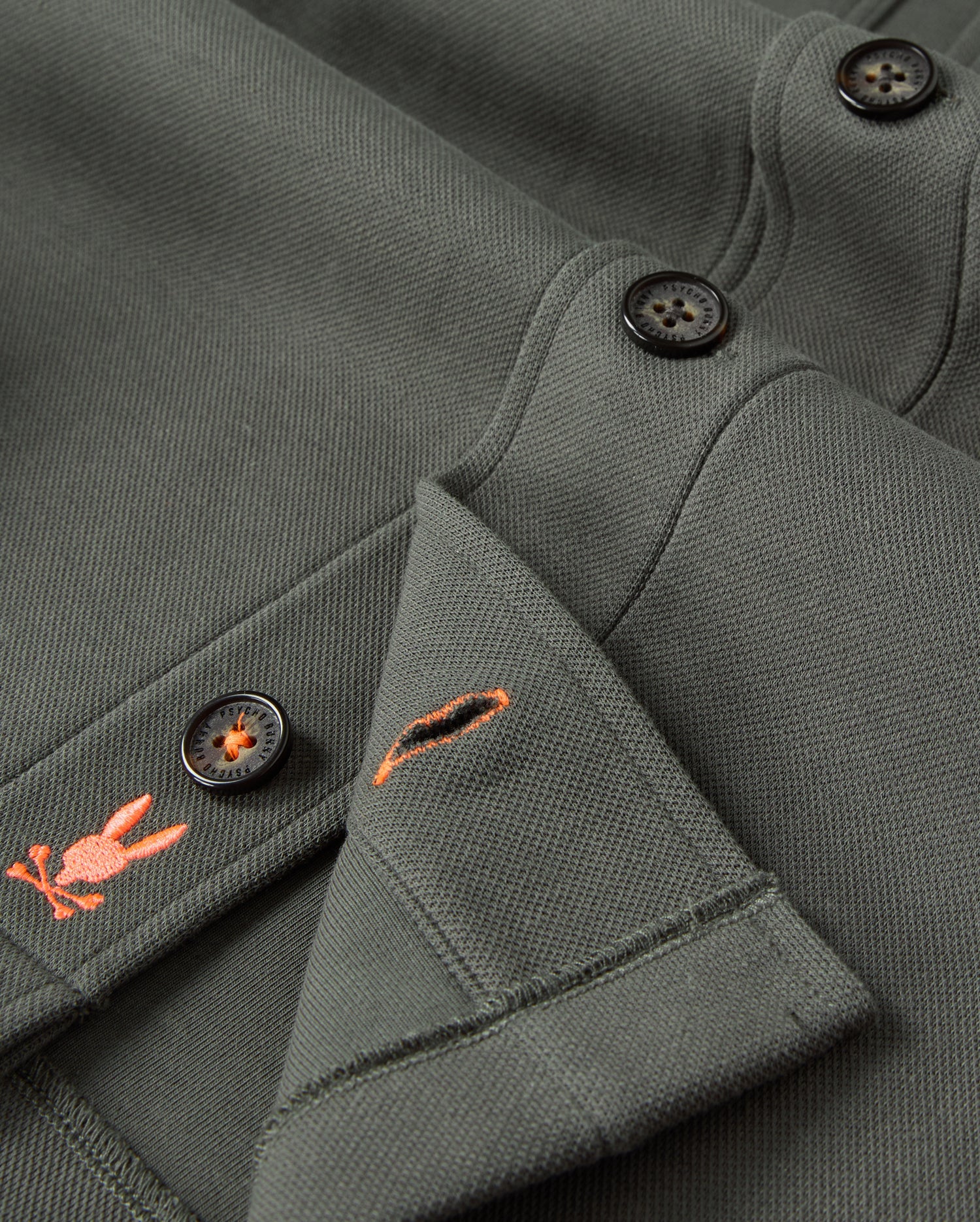 Close-up of the dark green Psycho Bunny Men's Bedford Shirt Jacket (B6J290B200) with black buttons. This cotton-blend piece features small embroidered designs of a carrot and a rabbit's head with crossbones in orange near the buttons, adding an edgy urban vibe.