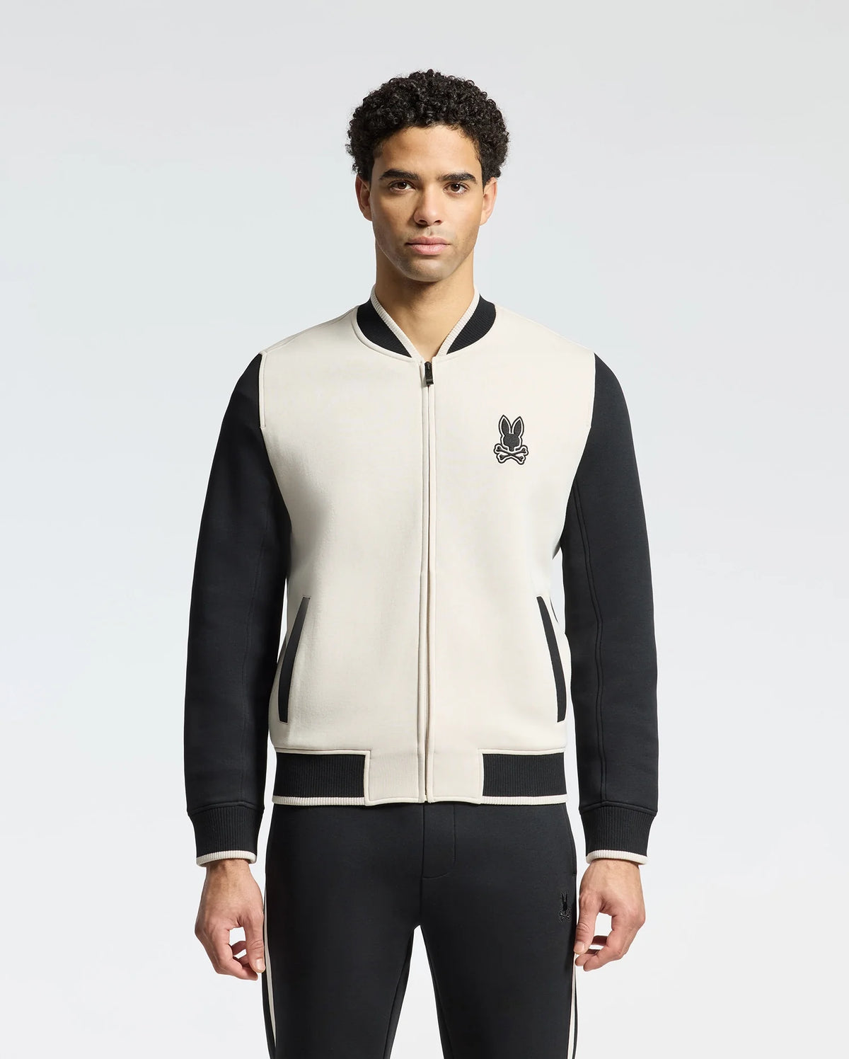 A man with curly hair models the Psycho Bunny MENS CRETE COLOR BLOCK BOMBER (B6J421F200), a light jacket with black sleeves and accents, featuring a small black embroidered logo on the chest. He pairs it with black pants, set against a plain light gray backdrop—ideal for transitional weather.