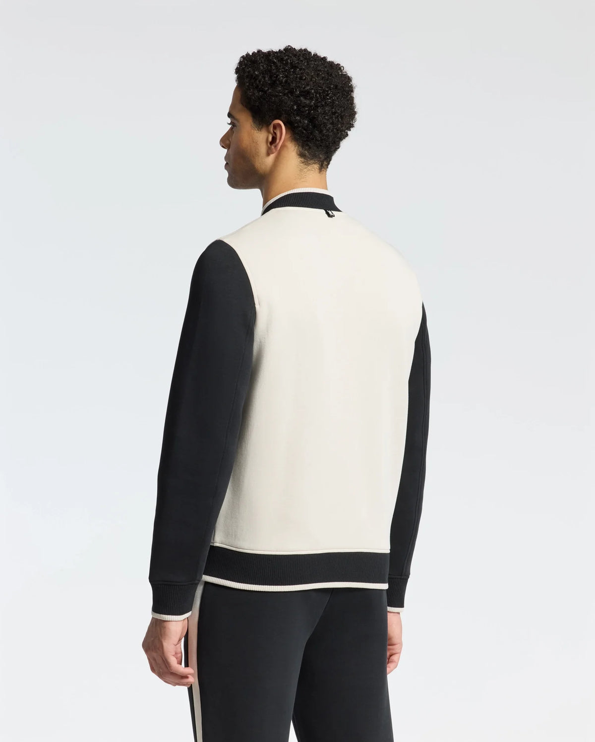A person with curly hair stands facing away, wearing Psycho Bunny's classic MENS CRETE COLOR BLOCK BOMBER - B6J421F200 in black and white, paired with matching track pants, set against a plain white background.