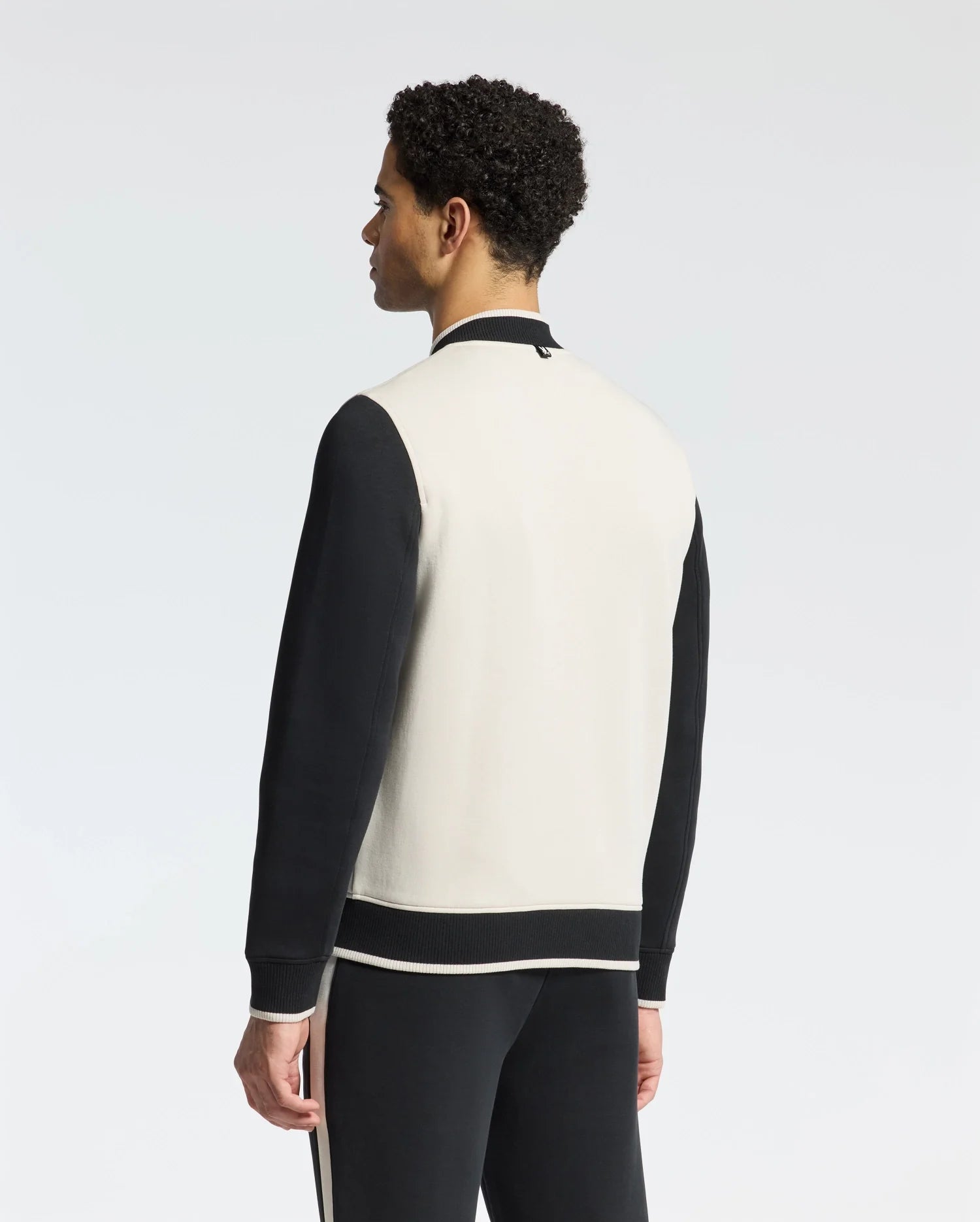 A person with curly hair stands facing away, wearing Psycho Bunny's classic MENS CRETE COLOR BLOCK BOMBER - B6J421F200 in black and white, paired with matching track pants, set against a plain white background.