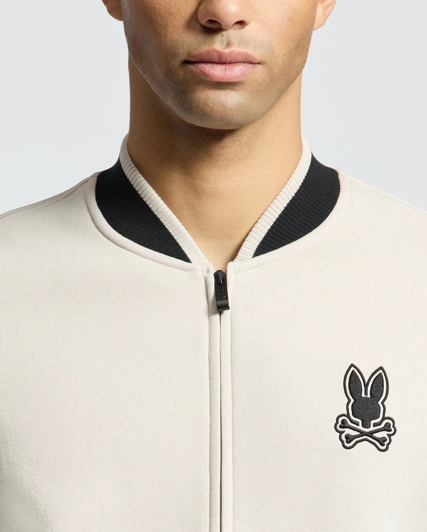 A person models the MENS CRETE COLOR BLOCK BOMBER - B6J421F200 by Psycho Bunny, showcasing its classic cut in beige with a black collar and a bunny head over crossbones emblem on the left side against a plain background.