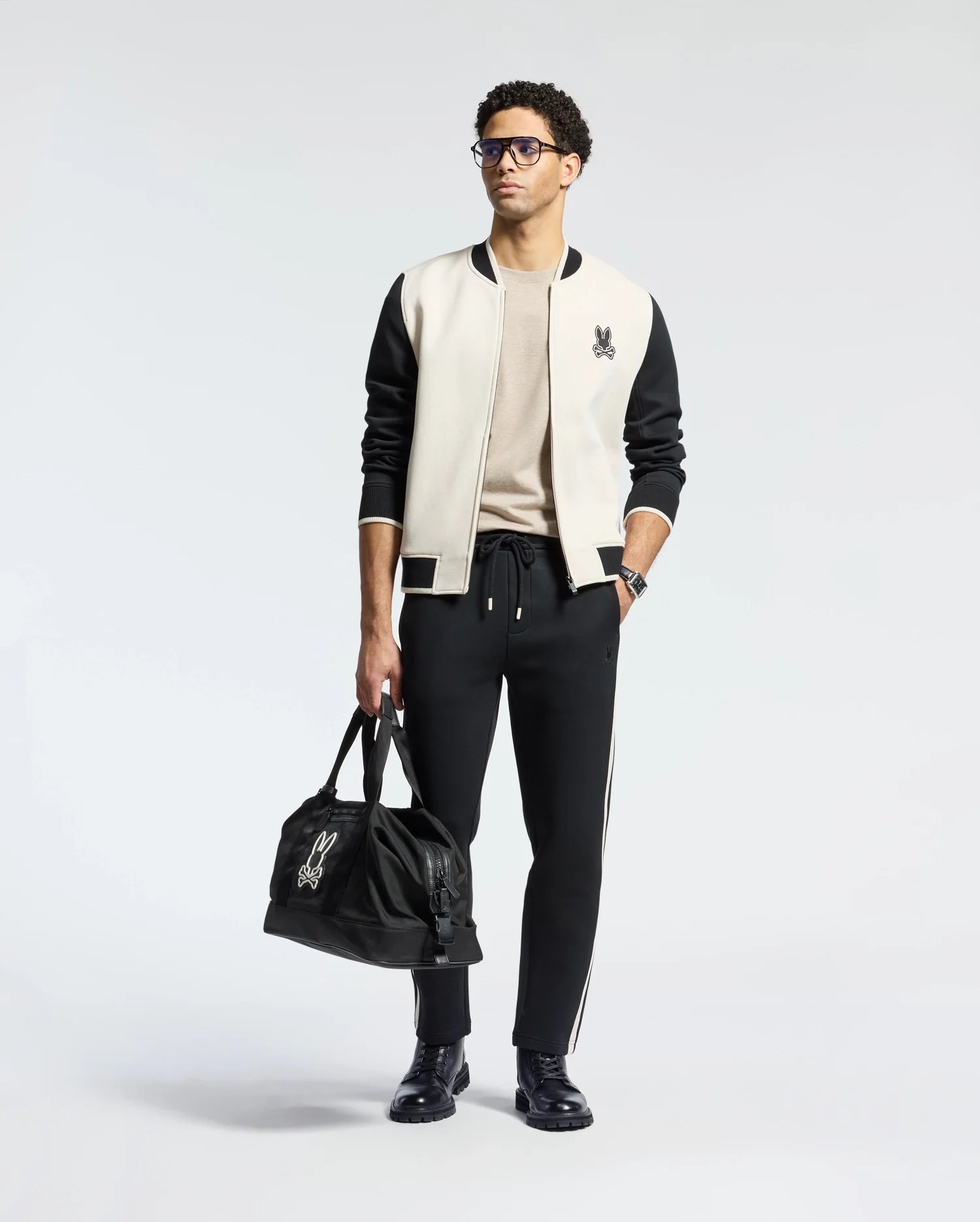 Man in glasses sports a classic look with the Psycho Bunny MENS CRETE COLOR BLOCK BOMBER jacket over a beige shirt, black pants, and shoes. He carries a logo-adorned black duffel bag on his left—a perfect transitional outfit against the white background.