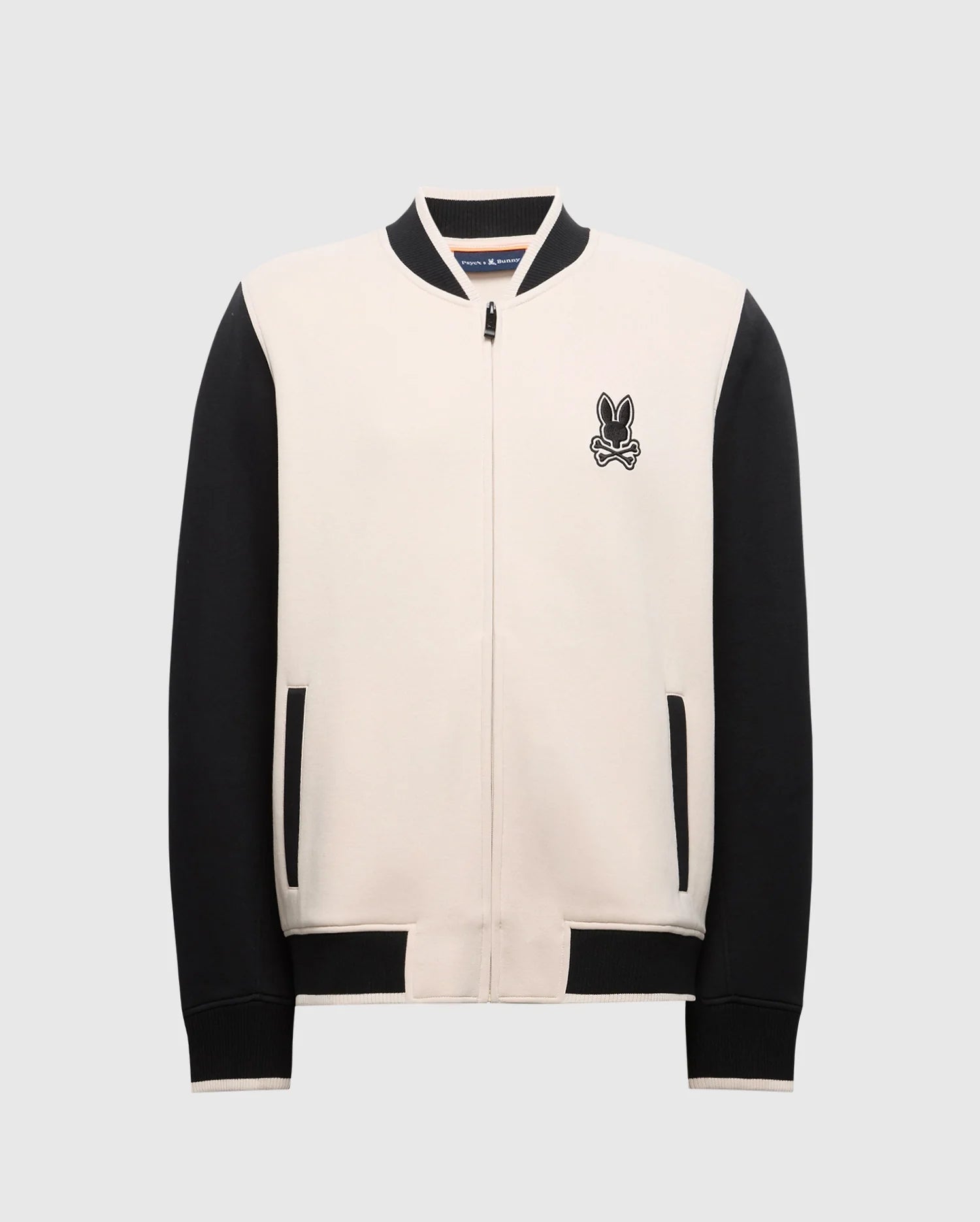 The Psycho Bunny MENS CRETE COLOR BLOCK BOMBER - B6J421F200 is a varsity-style jacket with a beige front and black sleeves. It includes a zipper closure, black pockets, an embroidered bunny logo on the left chest, and features black ribbed detailing on the collar, cuffs, and hem. Ideal for transitional wear.