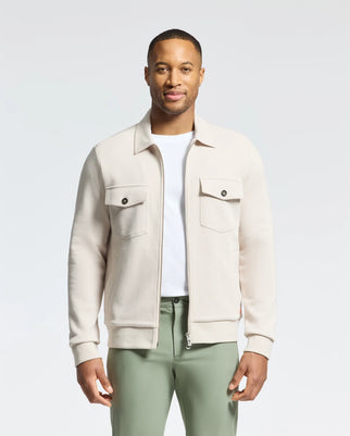A man wears the cream MENS THE OAKS KNIT BOMBER SHACKET from Psycho Bunny, featuring two front pockets and crafted from a premium cotton blend. He pairs it with a sleek white t-shirt and green pants, standing against a plain light background with a timeless look.