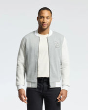 A man is wearing the Psycho Bunny MENS HYDRA COLOR BLOCK KNIT BOMBER - B6J455F200 in light gray and white premium cotton-blend with an embroidered Bunny "B" logo on the chest over a white shirt, paired with black pants. He stands against a plain white background.