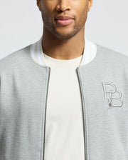 A person dons the Psycho Bunny MENS HYDRA COLOR BLOCK KNIT BOMBER in light gray, crafted from premium cotton-blend, with a white striped collar and zipper over a beige shirt. The embroidered "PB" logo with a small bunny adds charm. Focus is on the upper torso and part of the face with a neutral expression.