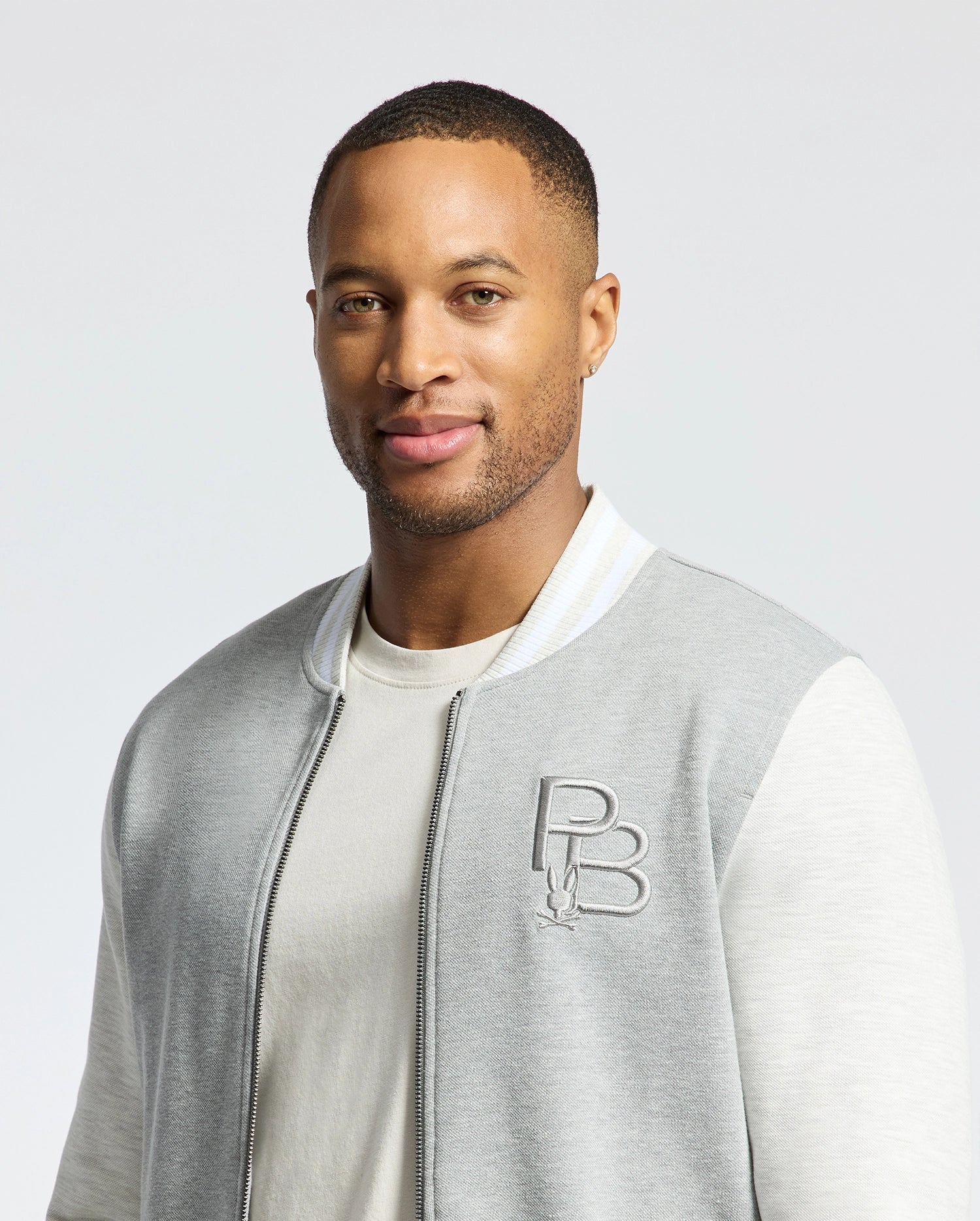 Someone is wearing a Psycho Bunny MENS HYDRA COLOR BLOCK KNIT BOMBER - B6J455F200 in light gray and white with an embroidered Bunny logo over a premium cotton-blend T-shirt. The background is plain white.