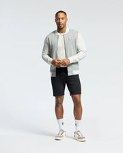 Against a plain background, a person wears Psycho Bunny's MENS HYDRA COLOR BLOCK KNIT BOMBER - B6J455F200 in light gray over a white shirt, paired with black shorts and white logo socks. Light brown sneakers complete the look. With a short beard and neutral expression, they exude laid-back style.