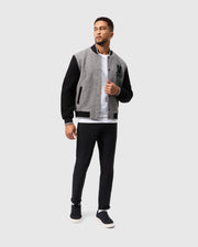 A man stands against a plain background wearing the Psycho Bunny MENS TOM WOOL BOMBER JACKET - B6J744D200, featuring stylish grey and black sections. He pairs it with a classic white t-shirt and black pants, completing the look with black sneakers. With one hand in his jacket pocket, he glances slightly to his left.