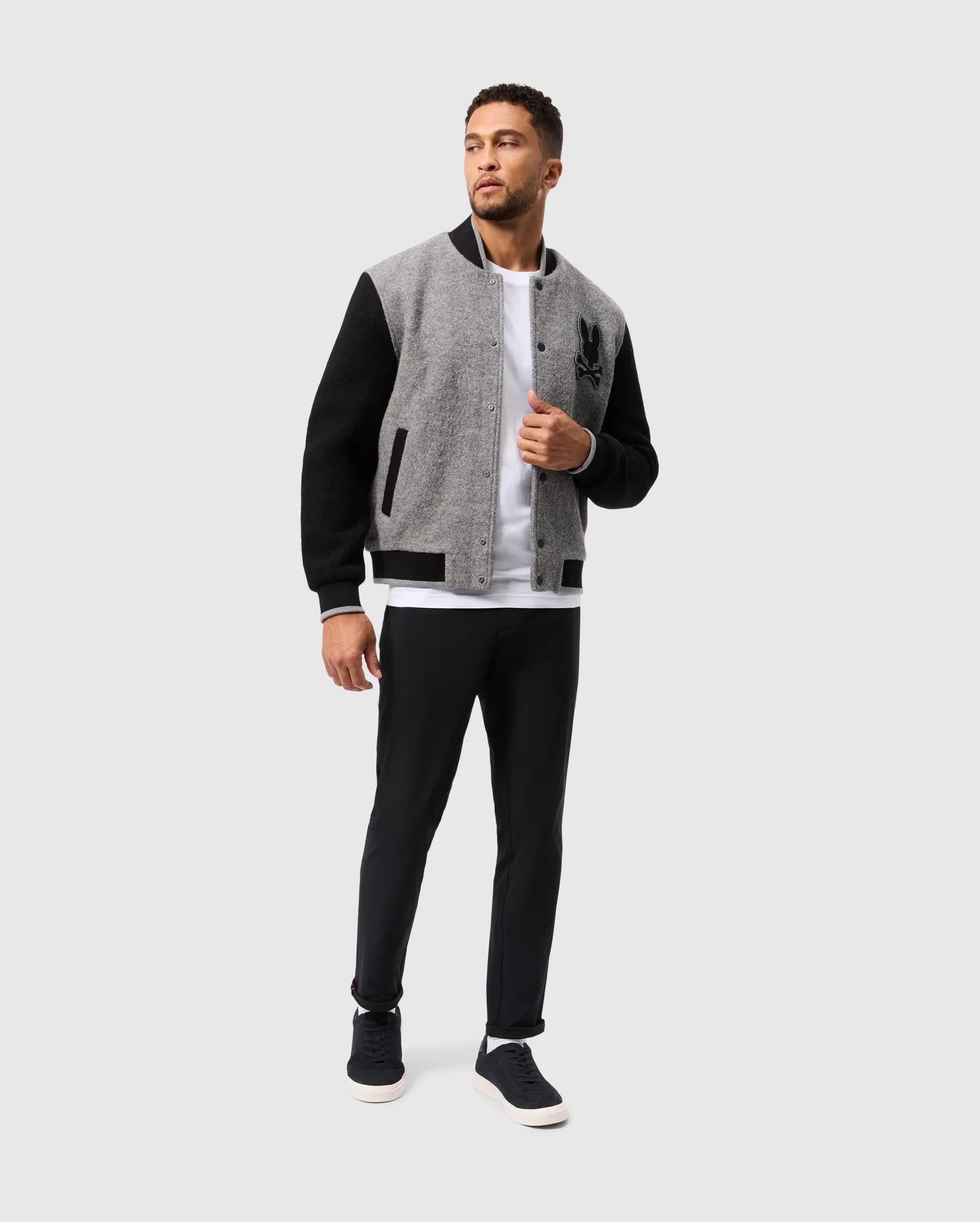 A man stands against a plain background wearing the Psycho Bunny MENS TOM WOOL BOMBER JACKET - B6J744D200, featuring stylish grey and black sections. He pairs it with a classic white t-shirt and black pants, completing the look with black sneakers. With one hand in his jacket pocket, he glances slightly to his left.