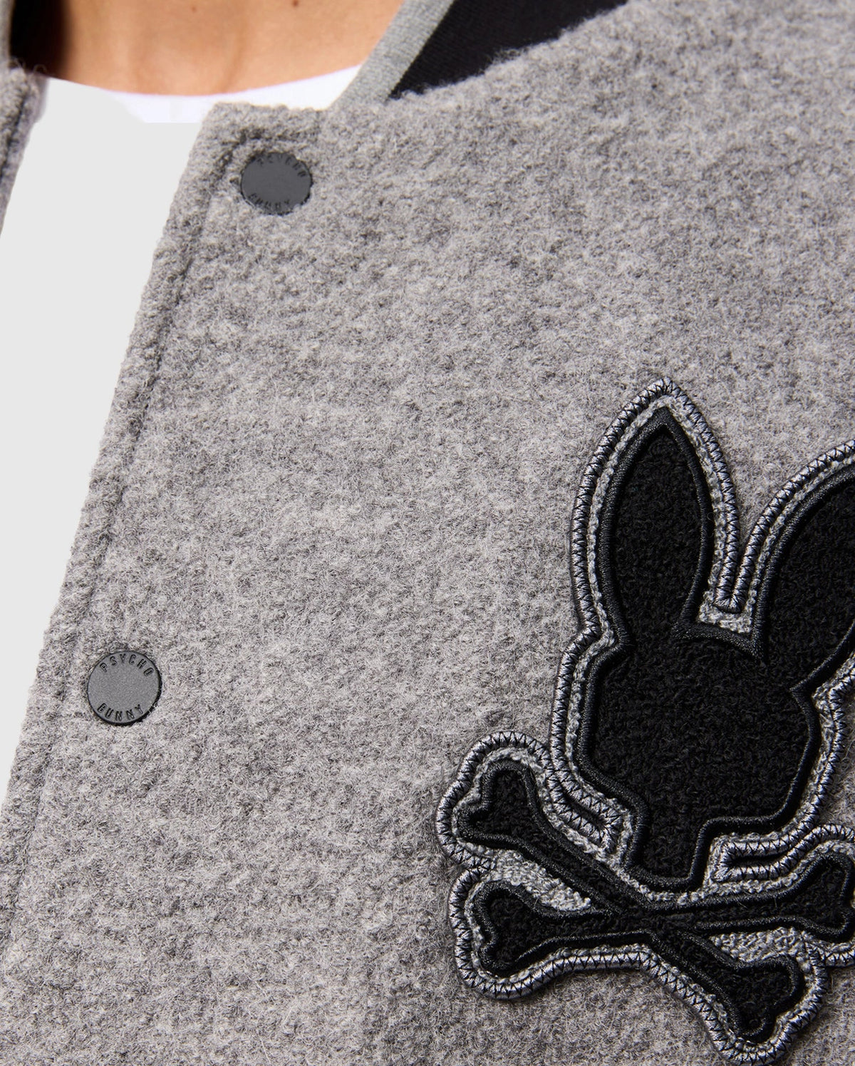 Close-up of the Psycho Bunny MENS TOM WOOL BOMBER JACKET - B6J744D200 in gray, featuring a black and gray embroidered patch with a rabbit head design and crossed bones beneath it. The premium fabric has a textured appearance, with part of a white shirt visible at the neck.