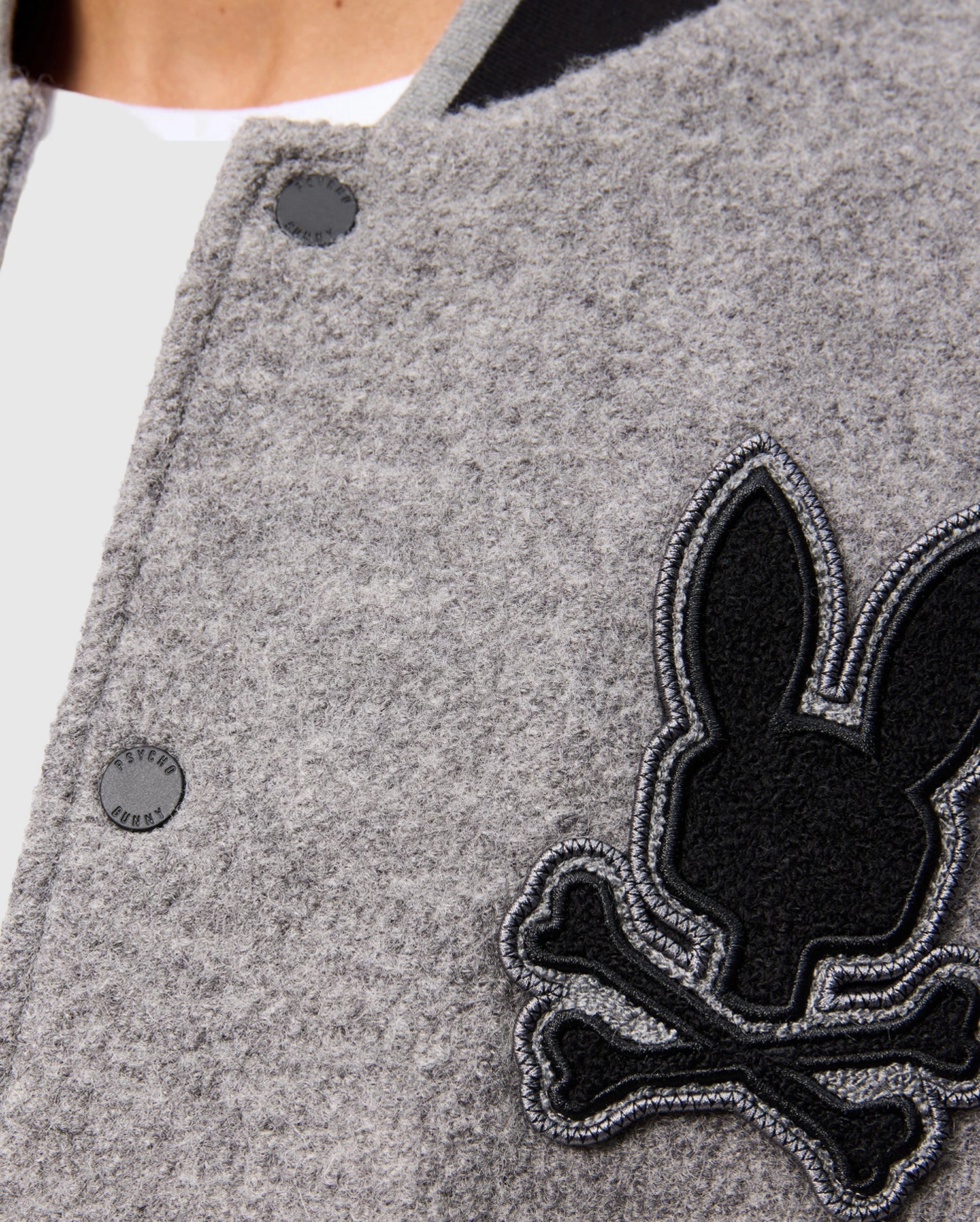 Close-up of the Psycho Bunny MENS TOM WOOL BOMBER JACKET - B6J744D200 in gray, featuring a black and gray embroidered patch with a rabbit head design and crossed bones beneath it. The premium fabric has a textured appearance, with part of a white shirt visible at the neck.