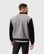A man is pictured from behind wearing the Psycho Bunny MENS TOM WOOL BOMBER JACKET - B6J744D200, which features a small chenille embroidered patch on the upper back. The jacket's sleeves and collar are black, while its body is crafted from gray virgin wool. He has short, curly hair and stands against a plain white background.