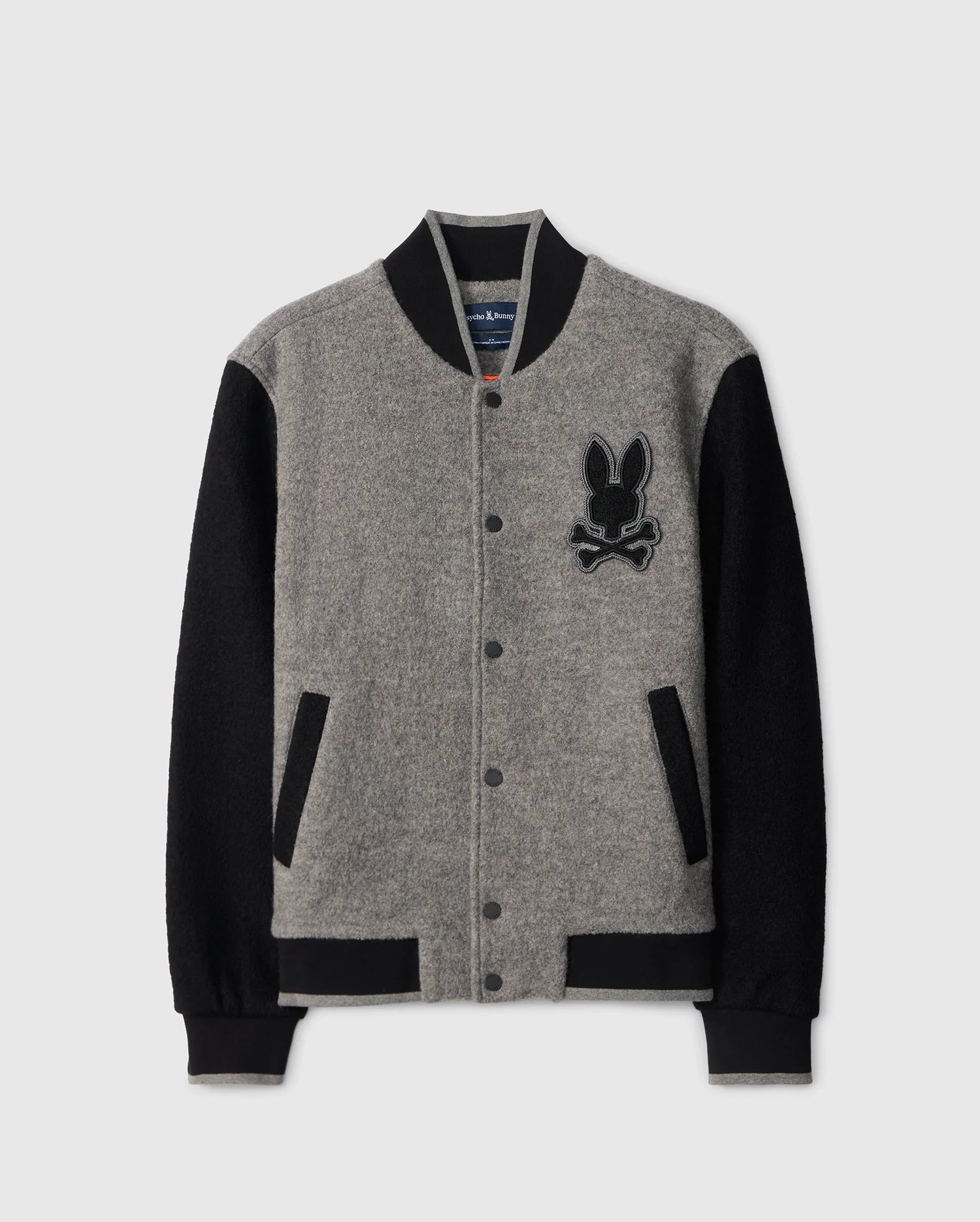 The MENS TOM WOOL BOMBER JACKET - B6J744D200 by Psycho Bunny is a stylish option with a grey body and black sleeves. It features black ribbed collar, cuffs, and hem, as well as a black embroidered patch on the left chest depicting a cartoonish rabbit's head with crossbones.