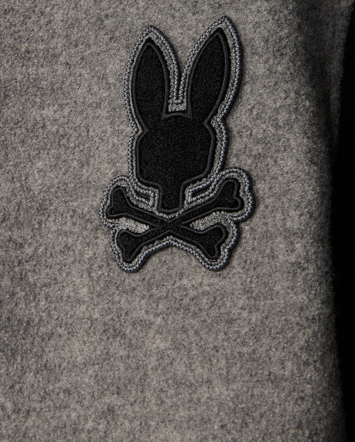 A close-up view of the grey fabric on the Psycho Bunny MENS TOM WOOL BOMBER JACKET - B6J744D200, featuring an embroidered black and grey chenille patch depicting a stylized bunny head above two crossed bones.