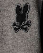A close-up view of the grey fabric on the Psycho Bunny MENS TOM WOOL BOMBER JACKET - B6J744D200, featuring an embroidered black and grey chenille patch depicting a stylized bunny head above two crossed bones.