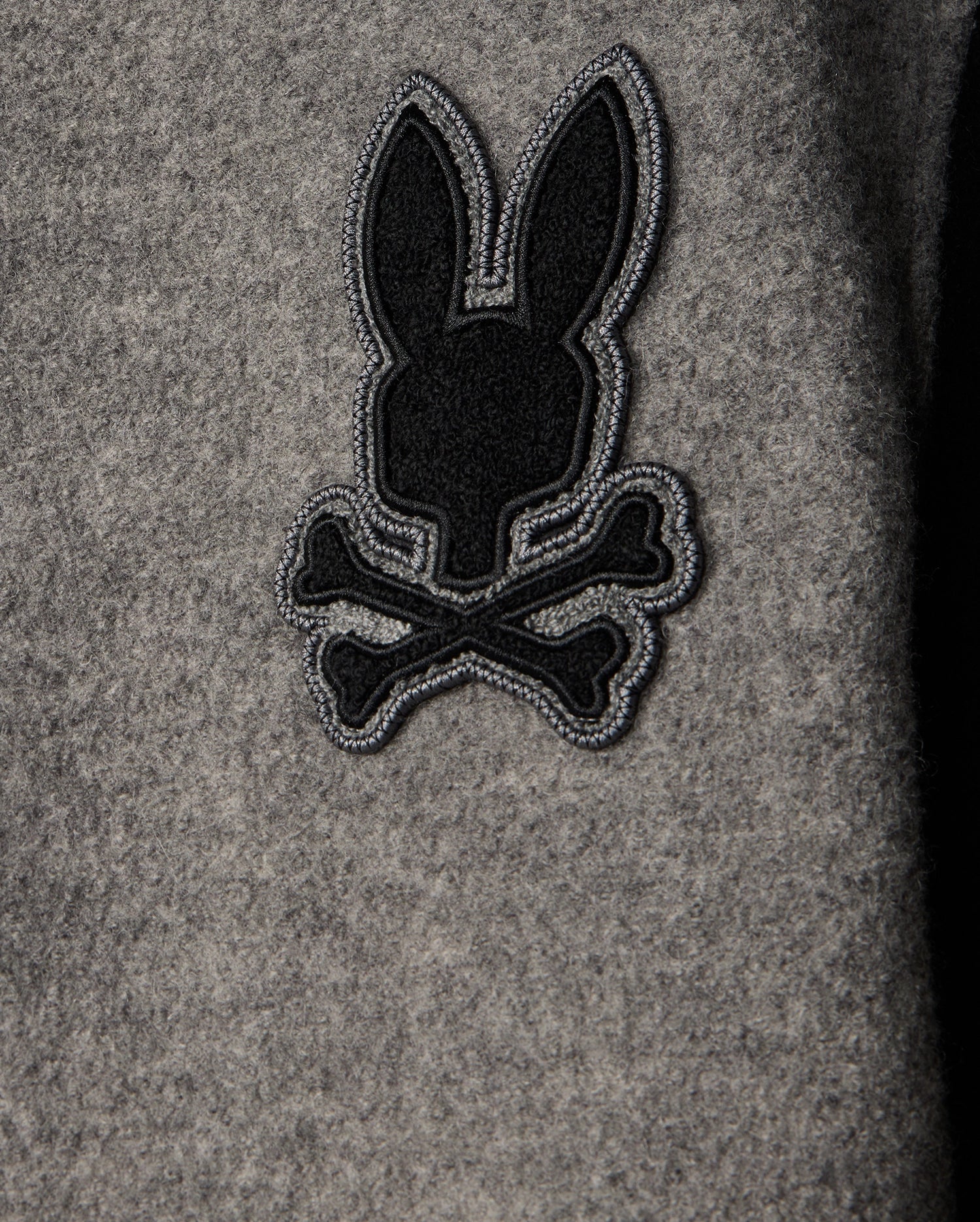 A close-up view of the grey fabric on the Psycho Bunny MENS TOM WOOL BOMBER JACKET - B6J744D200, featuring an embroidered black and grey chenille patch depicting a stylized bunny head above two crossed bones.