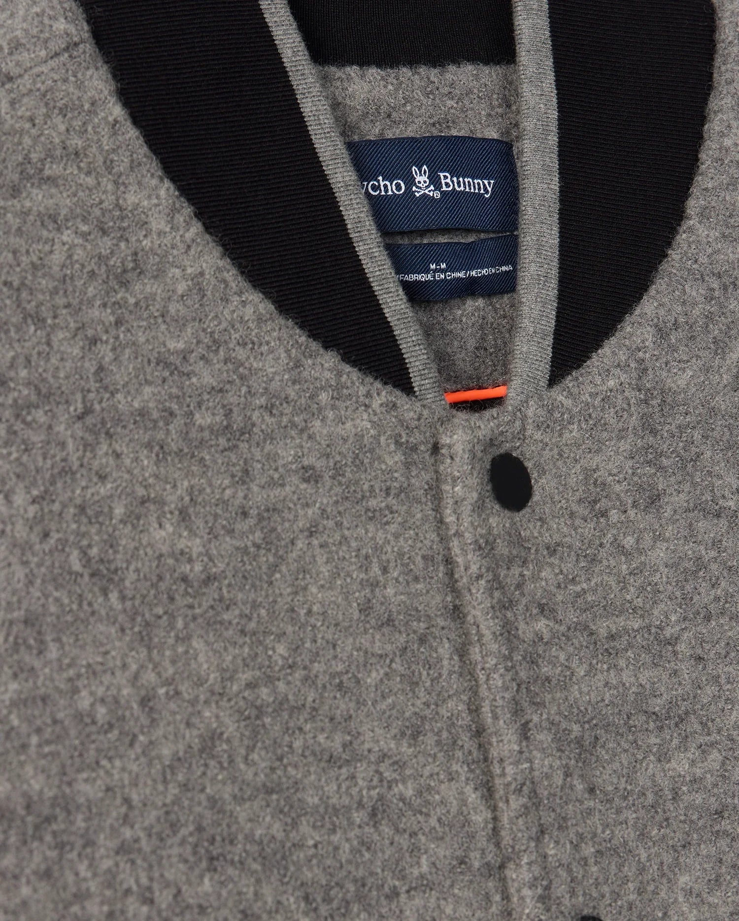 Close-up of a Men's Tom Wool Bomber Jacket (B6J744D200) by Psycho Bunny, featuring a gray wool body with a black collar and single black button. The inside label reads “Psycho Bunny” with a small chenille Bunny patch above it. The seam of the button placket and the top of the collar are visible, highlighting the fine craftsmanship using virgin wool.