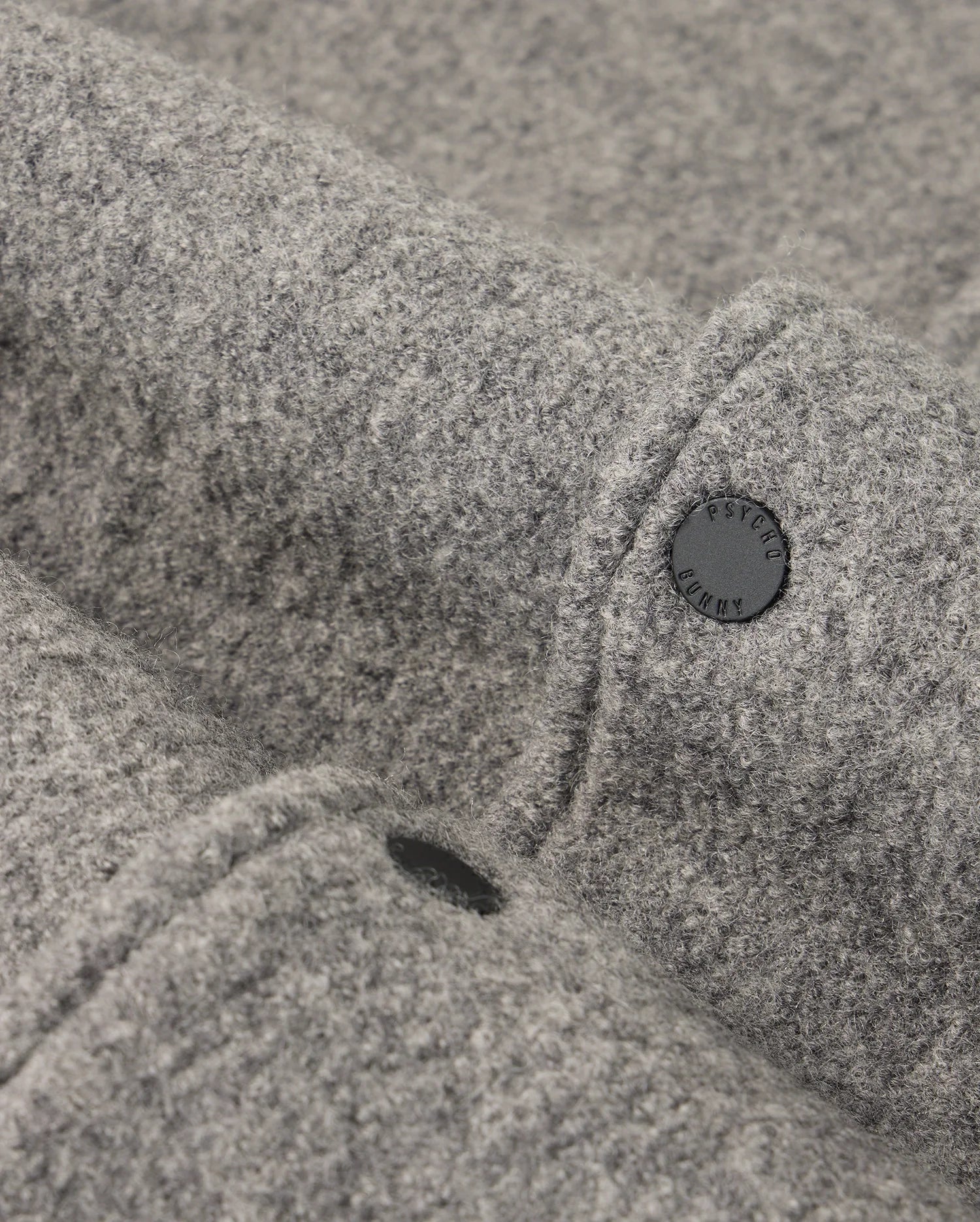 Close-up of a gray fabric texture showcasing the soft, felt-like material of the MENS TOM WOOL BOMBER JACKET - B6J744D200 by Psycho Bunny. Two black buttons are visible, partially embedded in the premium wool jacket, with one clearly displaying an engraved word in uppercase letters. The surface appears smooth and cozy.
