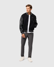 A man wearing the Psycho Bunny MENS BRAYDEN WOOL LEATHER VARSITY BOMBER - B6J876D200, featuring leather sleeves, pairs it with a white t-shirt, gray pants, and white sneakers as he stands against a plain background. With his hands in his jacket pockets, he gazes to the side.