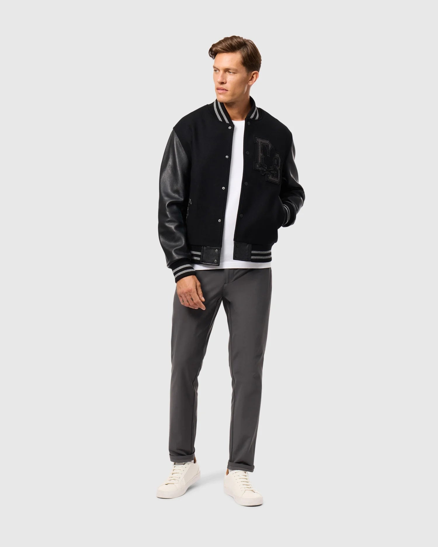 A man wearing the Psycho Bunny MENS BRAYDEN WOOL LEATHER VARSITY BOMBER - B6J876D200, featuring leather sleeves, pairs it with a white t-shirt, gray pants, and white sneakers as he stands against a plain background. With his hands in his jacket pockets, he gazes to the side.