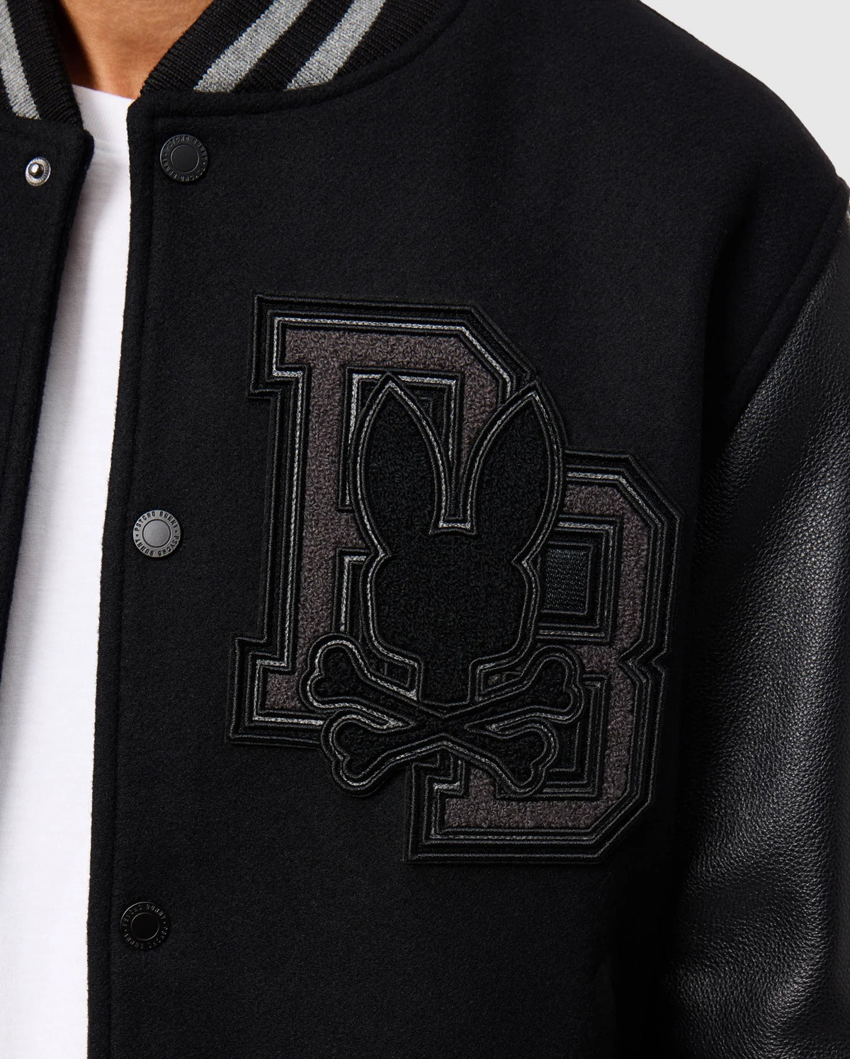 Close-up of the Psycho Bunny Men's Brayden Wool Leather Varsity Bomber (B6J876D200) in black, showcasing an embroidered design featuring large overlapping "DB" letters with a bunny head and crossed bones on the left chest. This wool-blend jacket includes snap buttons, leather sleeves, and a striped collar.