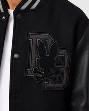 Close-up of the Psycho Bunny Men's Brayden Wool Leather Varsity Bomber (B6J876D200) in black, showcasing an embroidered design featuring large overlapping "DB" letters with a bunny head and crossed bones on the left chest. This wool-blend jacket includes snap buttons, leather sleeves, and a striped collar.