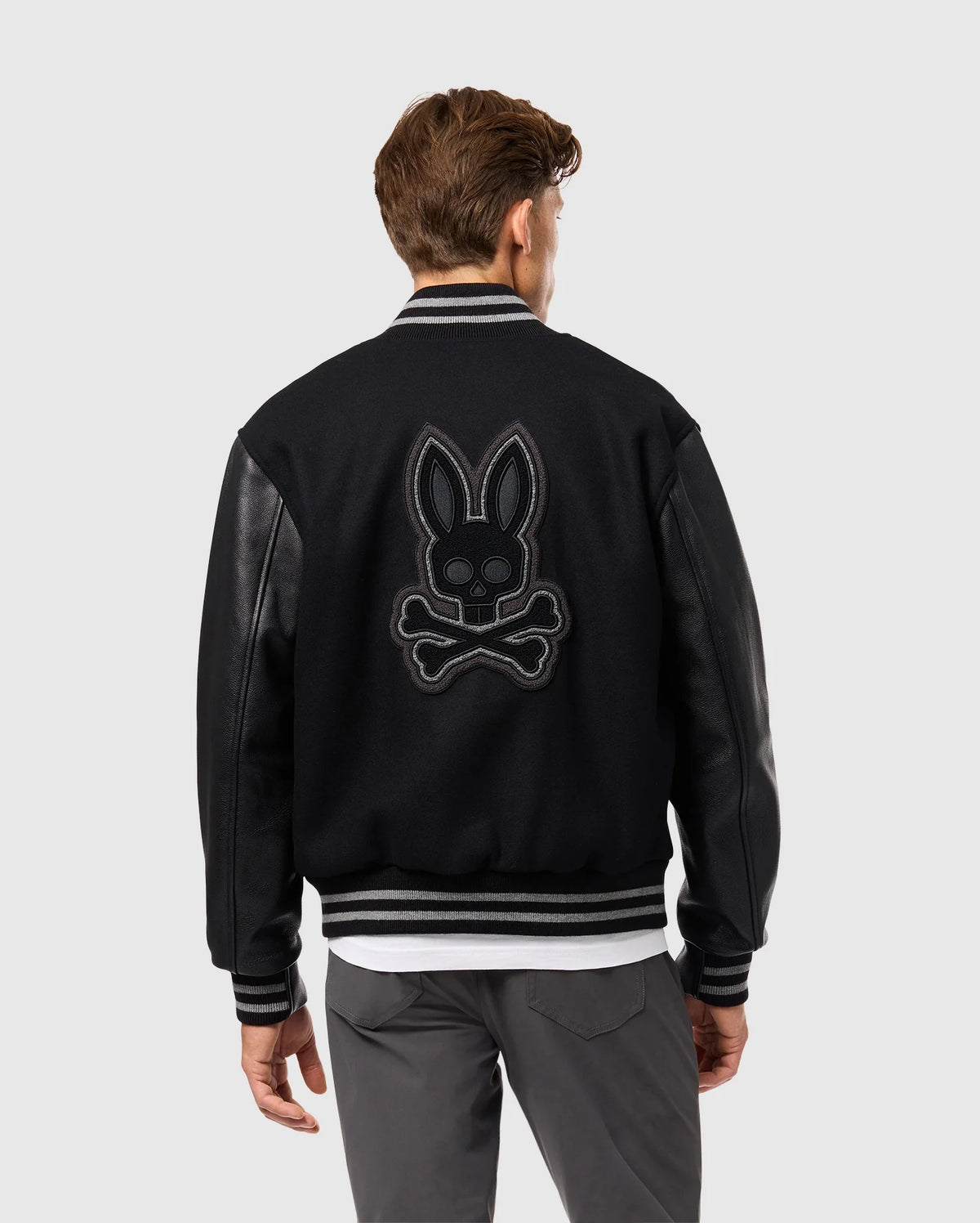 The individual sports the Psycho Bunny MENS BRAYDEN WOOL LEATHER VARSITY BOMBER - B6J876D200, featuring sleek leather sleeves. The back displays a striking skull-and-crossbones motif adorned with rabbit ears. With short brown hair, they're turned away, stylishly pairing the jacket with grey pants and a white shirt beneath.