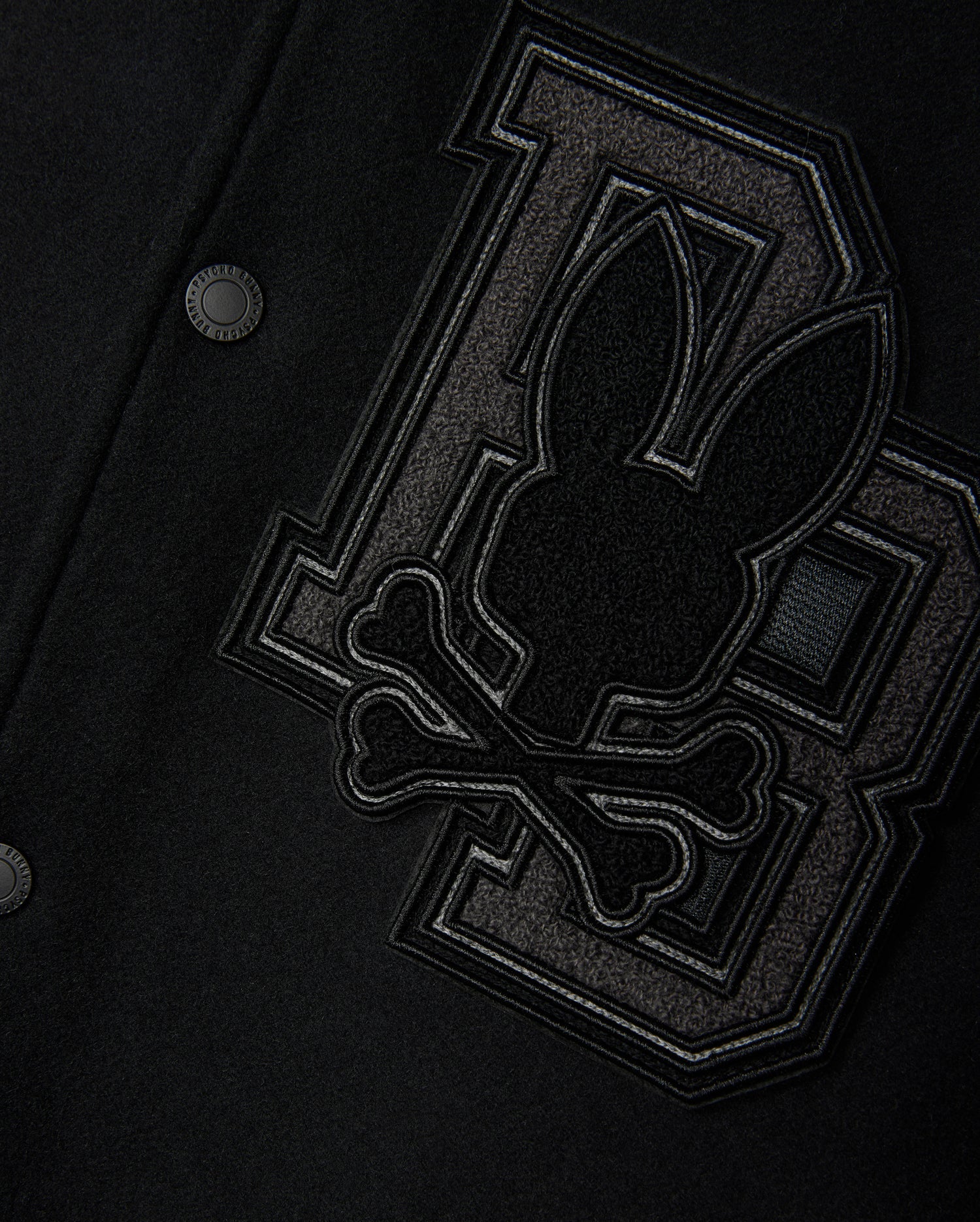 Close-up of the Psycho Bunny Men's Brayden Wool Leather Varsity Bomber - B6J876D200 in black, featuring leather sleeves and a prominent patch. The patch showcases a stylized rabbit head above crossbones, positioned against two large interlocking "BB" letters crafted from textured fabric. This wool-blend jacket is finished with metal snap buttons.