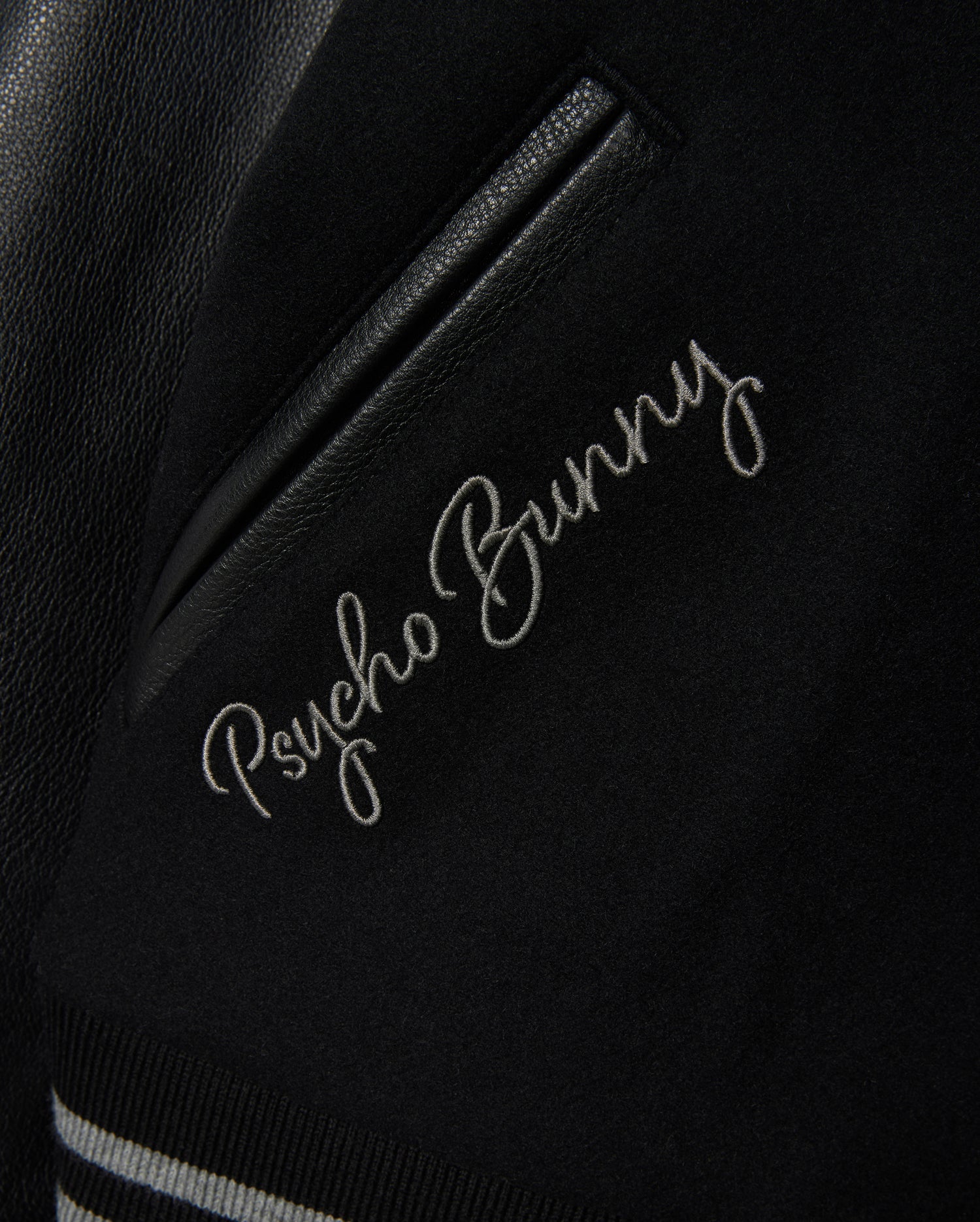 Detailed view of the MENS BRAYDEN WOOL LEATHER VARSITY BOMBER jacket, featuring "Psycho Bunny" elegantly embroidered in white cursive. This black bomber includes leather sleeves, a leather accent, and striped ribbed trim.