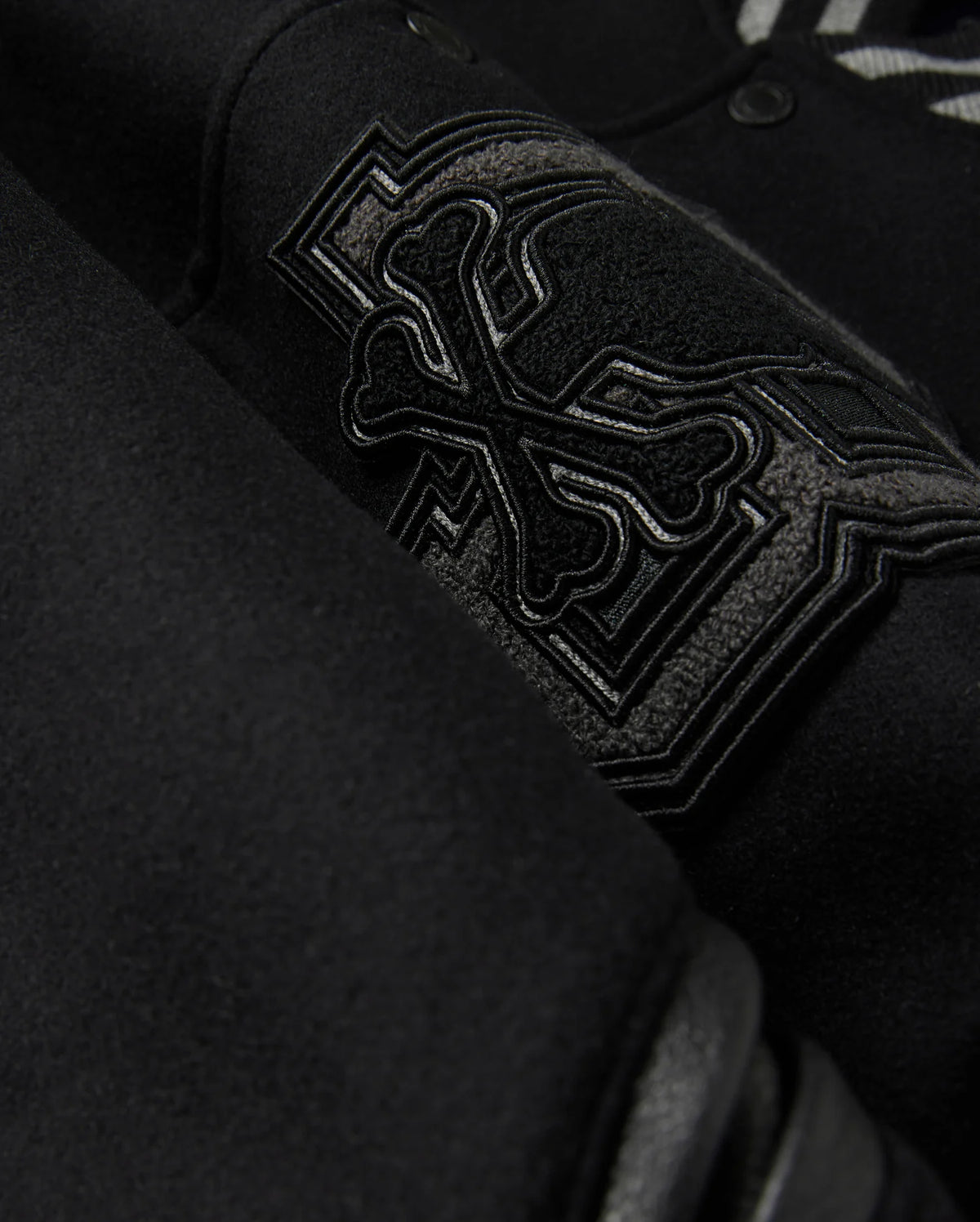 A detailed close-up of the MENS BRAYDEN WOOL LEATHER VARSITY BOMBER - B6J876D200 by Psycho Bunny showcases a black wool-blend fabric with an intricate embroidered design featuring crossbones and decorative elements. The raised and detailed stitching adds texture against the smooth background, reflecting the exceptional craftsmanship typical of a varsity bomber.