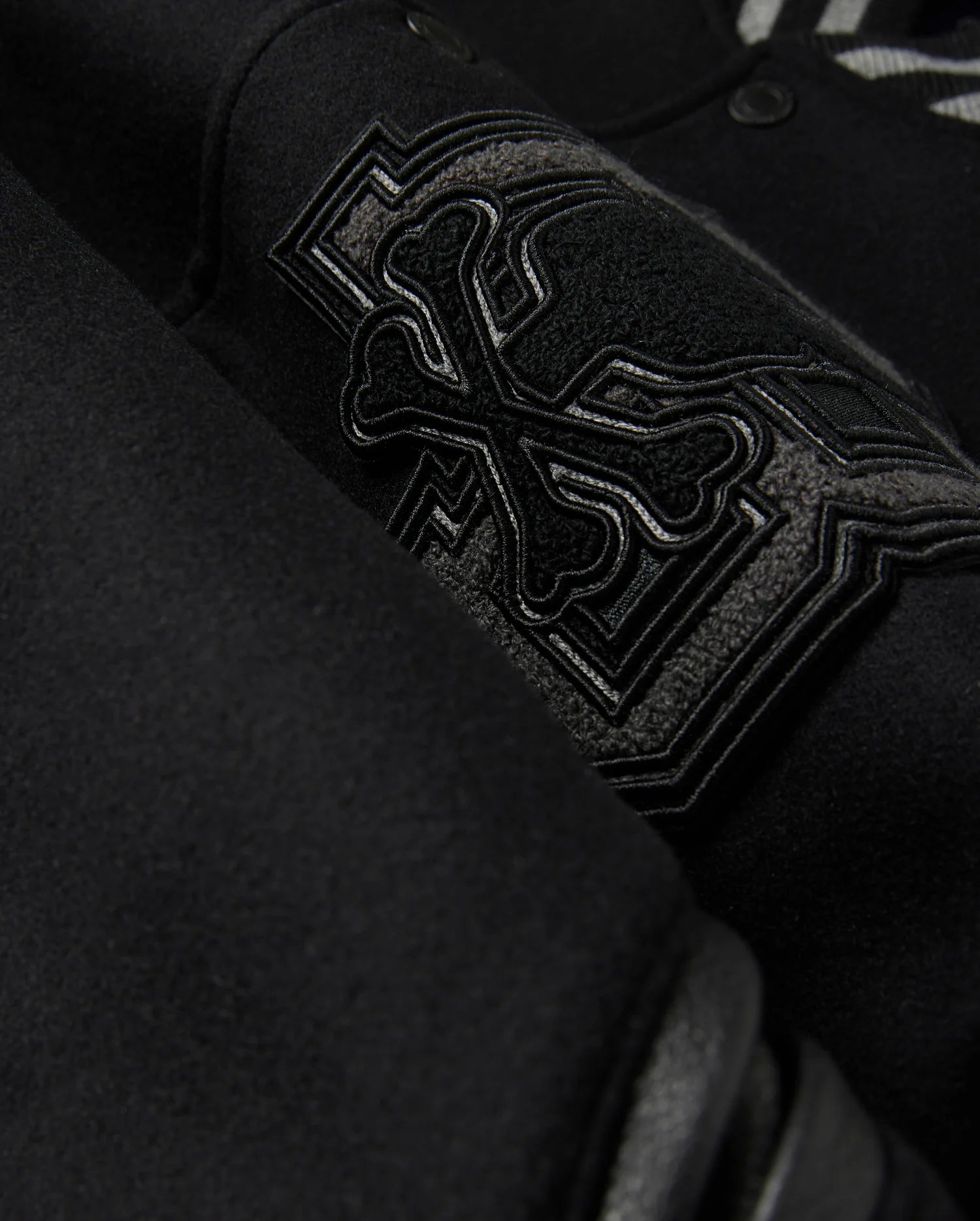 A detailed close-up of the MENS BRAYDEN WOOL LEATHER VARSITY BOMBER - B6J876D200 by Psycho Bunny showcases a black wool-blend fabric with an intricate embroidered design featuring crossbones and decorative elements. The raised and detailed stitching adds texture against the smooth background, reflecting the exceptional craftsmanship typical of a varsity bomber.