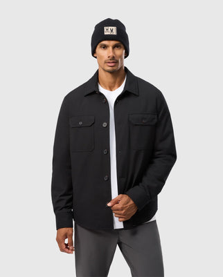A person is wearing a black beanie with a beige patch, a white T-shirt, and the Psycho Bunny MENS HILLCREST PADDED SHACKET (B6J936D200) with patch pockets. The individual is standing against a plain, light gray background and has one hand partially in their dark gray pants pocket.