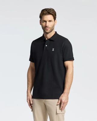 A man with short brown hair and a beard wears a Psycho Bunny Men's Classic Piqué Polo Shirt (B6K001ARPC) in black, made from 100% Pima cotton with mother-of-pearl buttons and a chest logo. He stands facing the camera with light beige pants against a plain, light-colored background.