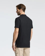 A man with short brown hair faces away, wearing a Psycho Bunny black Men's Classic Pique Polo Shirt (B6K001ARPC) made of 100% Pima cotton and beige pants. The polo features elegant mother-of-pearl buttons against a plain white background.
