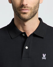 A person wears the Psycho Bunny Men's Classic Pique Polo Shirt (B6K001ARPC) made of 100% Pima cotton. The black polo features a small rabbit skull and crossbones logo, a classic collar, and mother-of-pearl buttons. The individual has facial hair against a plain background.