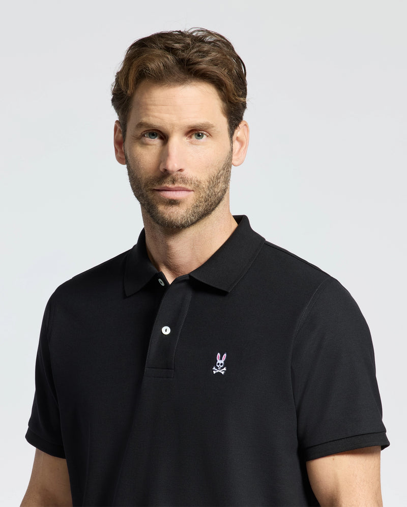 A man with short brown hair and a beard wears a Psycho Bunny Men's Classic Pique Polo Shirt - B6K001ARPC, crafted from 100% Pima cotton, featuring a small bunny logo and mother-of-pearl buttons. He stands against a plain light background, looking directly at the camera.