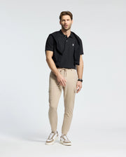 A man sports a Psycho Bunny MENS CLASSIC PIQUE POLO SHIRT - B6K001ARPC in black, paired with light tan joggers and white sneakers. A black sweater drapes over his shoulders, and a watch graces his wrist as he stands against a white background, looking straight ahead.