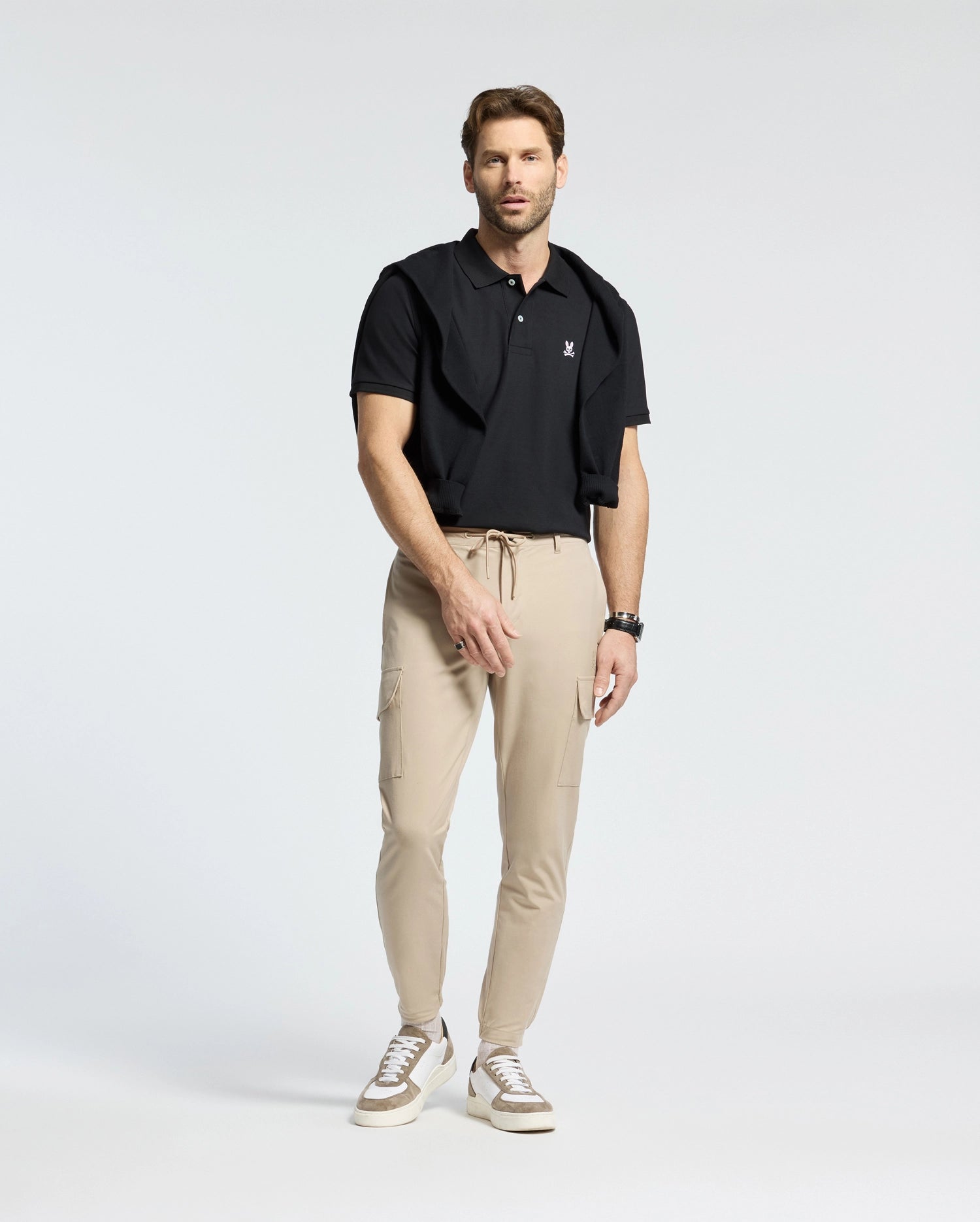 A man sports a Psycho Bunny MENS CLASSIC PIQUE POLO SHIRT - B6K001ARPC in black, paired with light tan joggers and white sneakers. A black sweater drapes over his shoulders, and a watch graces his wrist as he stands against a white background, looking straight ahead.