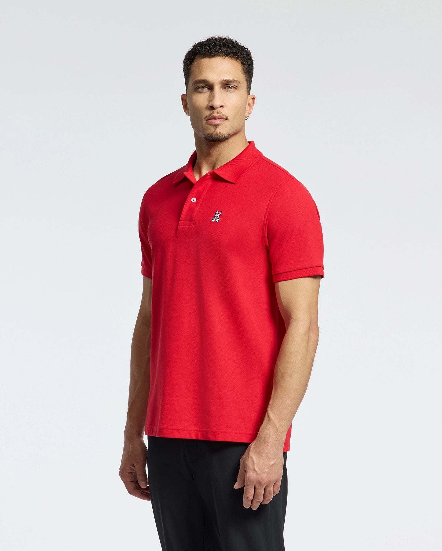 A man wearing a Psycho Bunny Men's Classic Pique Polo Shirt in red, made from soft Pima cotton with a small embroidered logo and elegant mother-of-pearl buttons, stands against a plain white background. He has short curly hair and pairs the polo with sleek black pants.