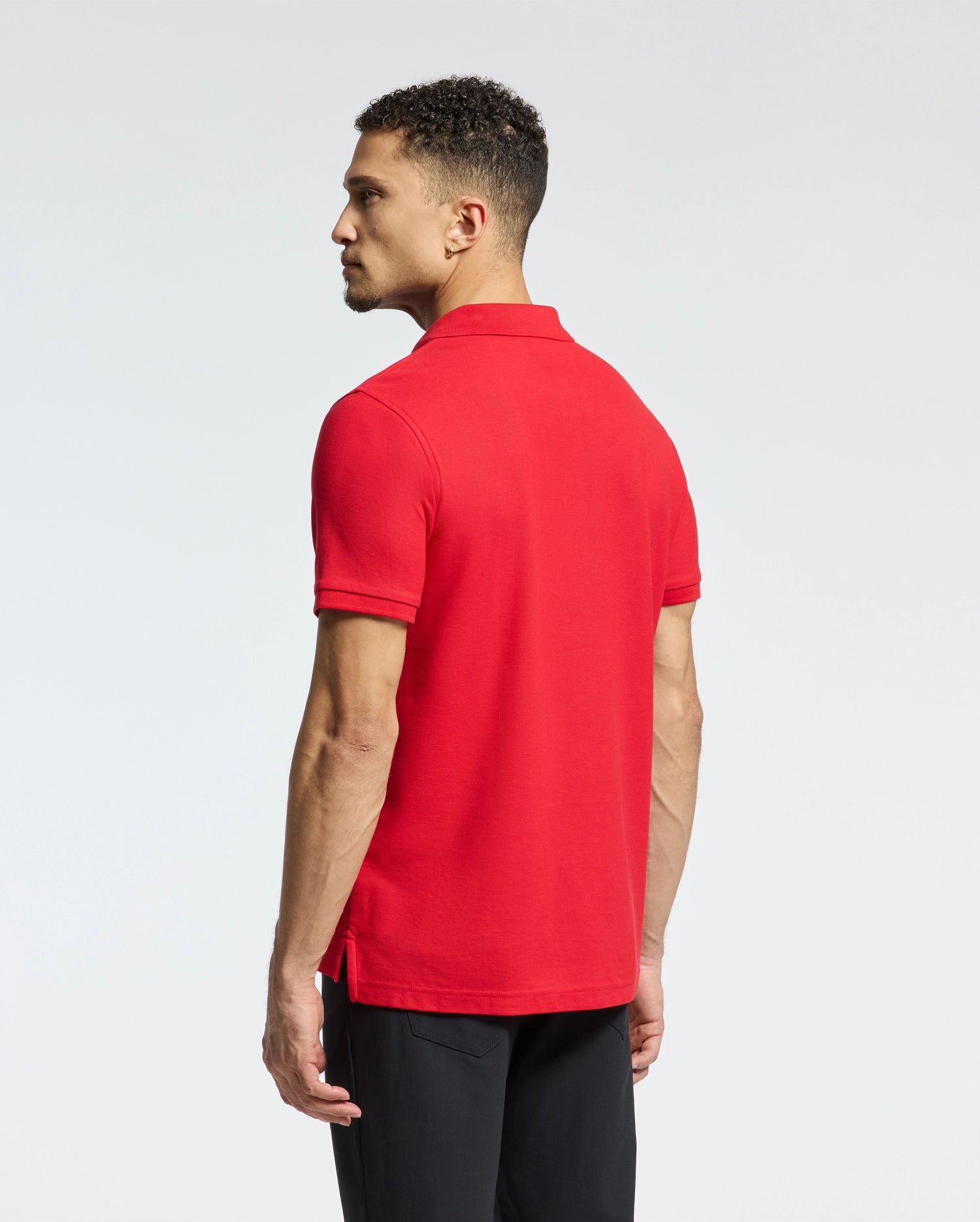 A man with short curly hair wears a Psycho Bunny Men's Classic Pique Polo Shirt, highlighted by mother-of-pearl buttons. The bright red shirt and black pants create a striking profile against the plain light background.