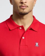 A person wearing a Psycho Bunny MENS CLASSIC PIQUE POLO SHIRT in rich red, featuring a small white bunny and crossbones logo on the chest. Crafted from soft Pima cotton, it has a collar with two elegant mother-of-pearl buttons.