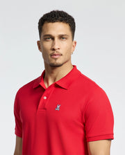 A person in a bright red Psycho Bunny MENS CLASSIC PIQUE POLO SHIRT - B6K001CRPC, crafted from soft Pima cotton and featuring a small bunny logo on the chest, stands against a plain backdrop. They have short, curly hair and an earring in one ear.