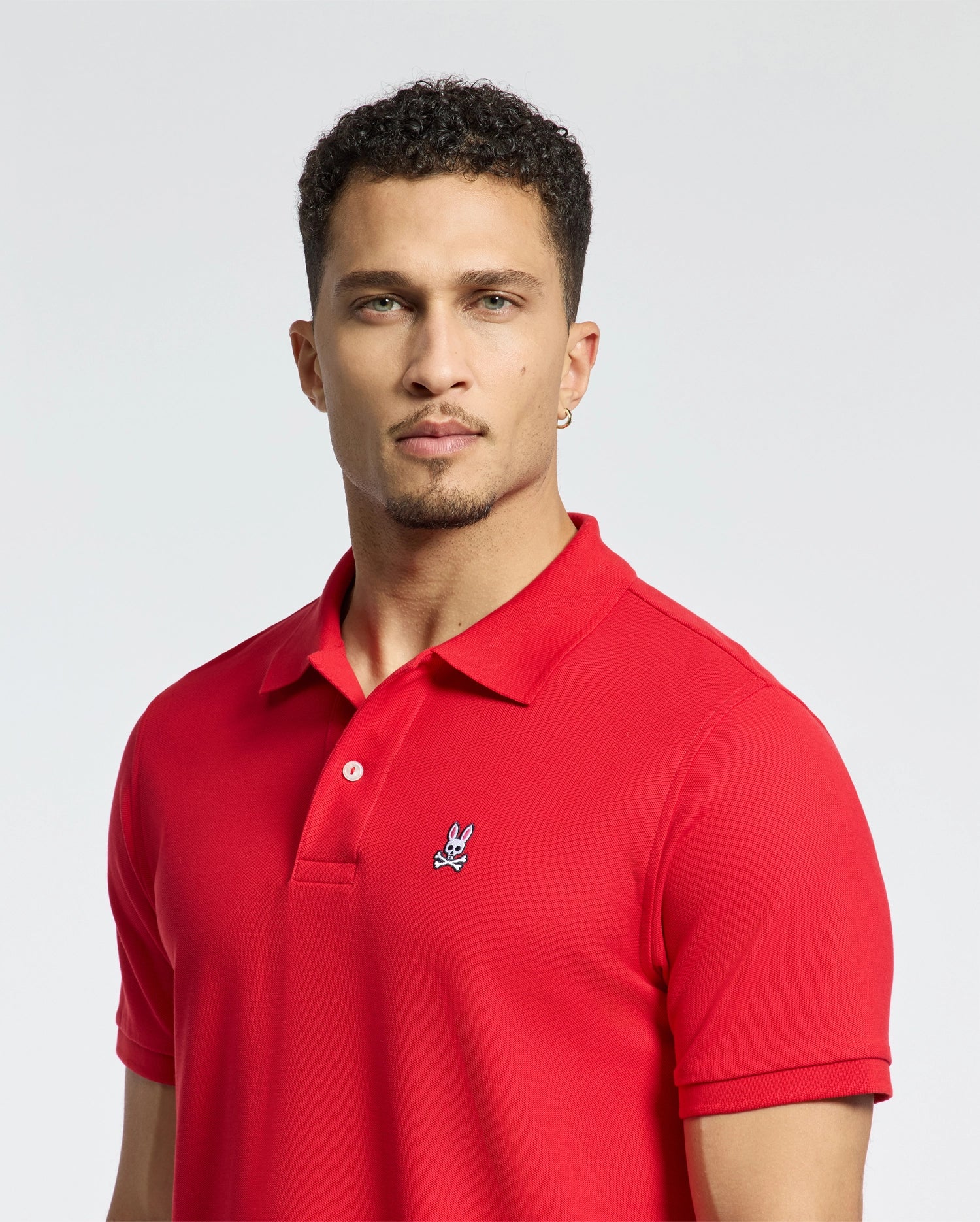 A person in a bright red Psycho Bunny MENS CLASSIC PIQUE POLO SHIRT - B6K001CRPC, crafted from soft Pima cotton and featuring a small bunny logo on the chest, stands against a plain backdrop. They have short, curly hair and an earring in one ear.