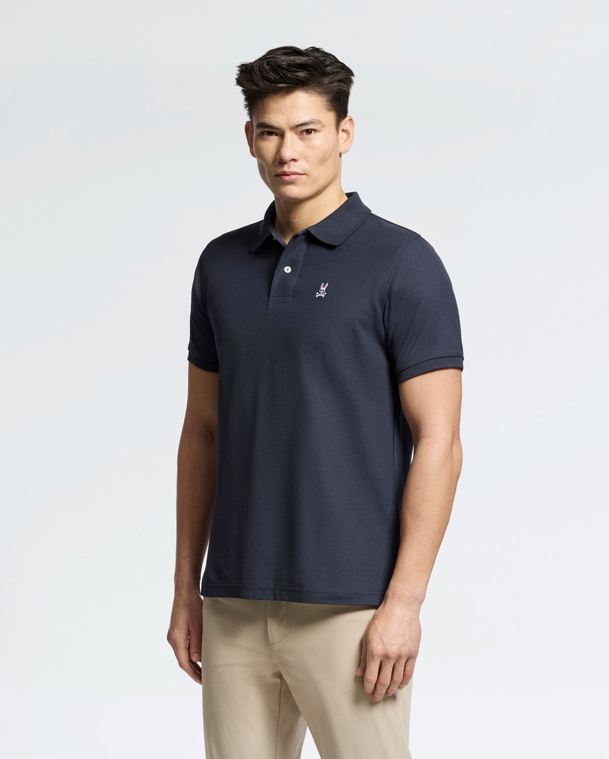 A man in a Psycho Bunny Men's Classic Pique Polo Shirt (B6K001ARPC) in navy, featuring 100% Pima cotton, an embroidered logo, and mother-of-pearl buttons, pairs it with beige pants against a plain light background.