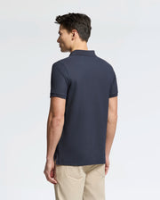 A person with short dark hair, wearing a Psycho Bunny Men's Classic Pique Polo Shirt (B6K001ARPC) in navy blue with mother-of-pearl buttons and beige pants, stands facing away against a white backdrop.