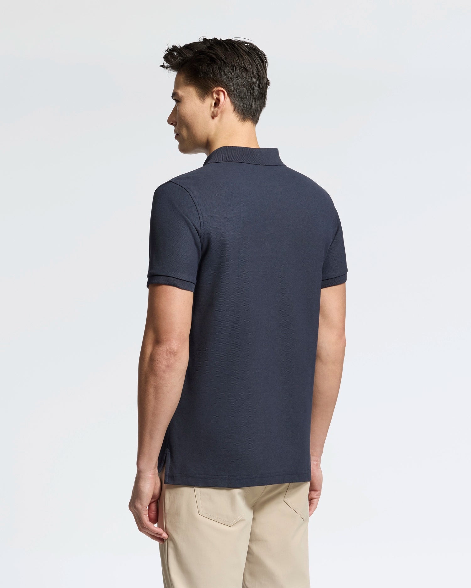 A person with short dark hair, wearing a Psycho Bunny Men's Classic Pique Polo Shirt (B6K001ARPC) in navy blue with mother-of-pearl buttons and beige pants, stands facing away against a white backdrop.