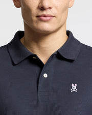 A person wears a Psycho Bunny Men's Classic Pique Polo Shirt (B6K001ARPC), made from 100% Pima cotton with mother-of-pearl buttons. The dark shirt features an embroidered pink-eared bunny logo with a skull and crossbones on the left chest against a plain white background.